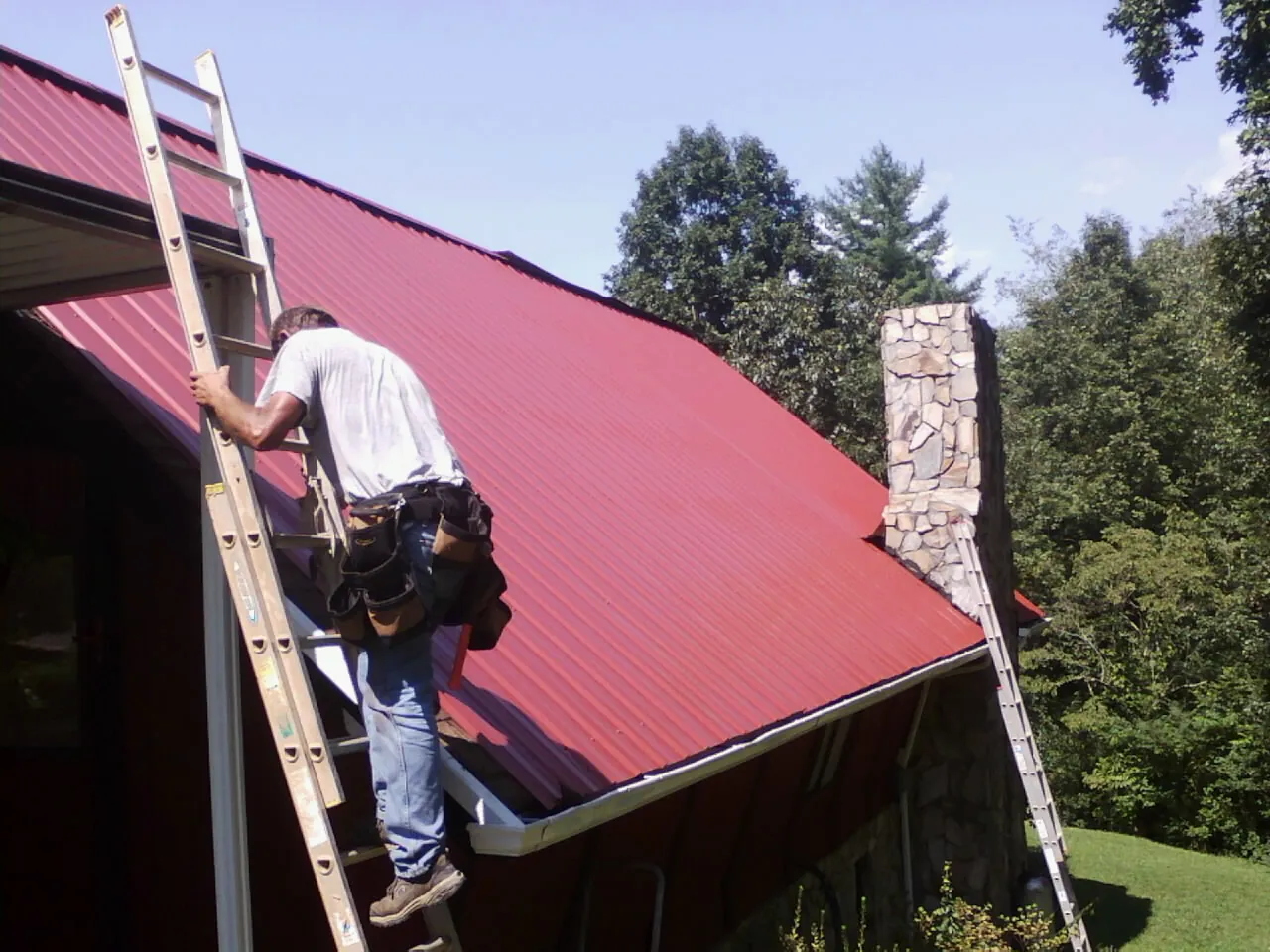 Roofer on a ladder beside a red metal roof and stone chimney — Soffit & Fascia Repair by Roofing Brothers in Detroit
